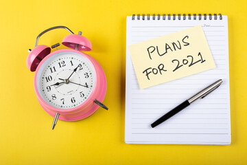 A table clock with Plans for 2021 words Written on a sticky note with notepad and other elements on a Yellow background