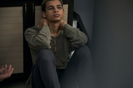 Staged photo illustrates problems and conflicts in gay couple relationships. Wistful swarthy man in gray cardigan and trousers is sitting near kitchen base cabinet. Other person's hand is in frame.
