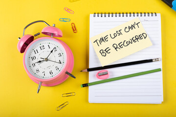 A table clock with Time lost can not be recovered words Written on a sticky note with notepad pencil and other elements on a Yellow background.