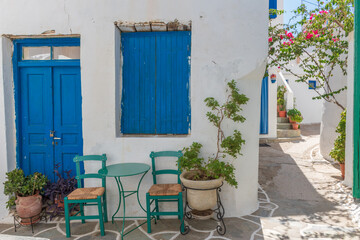 Fototapeta na wymiar Cycladitic alley with traditional green chairs and table, atypical facade of a house with a blue door and window and a blooming bougainvillea in Chora kythnos, cyclades, Greece.