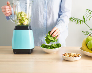 Raw foodism, green smoothie with banana and spinach. Woman putting spinach leaves to blender, dietary plant based mix with vegetables and fruits for vegan or vegetarian on table in home kitchen