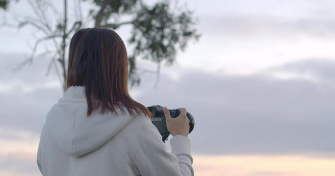 Rear view of woman photographer standing on the hill taking picture at the sunrise by using digital camera in slow motion shot. 