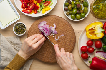 Vegetarian food. Chopping onion, cutting vegetables for greek salad horiatiki. Woman hands with knife on wooden cutting board slicing chop of vegetables. Homemade vegan food, top view