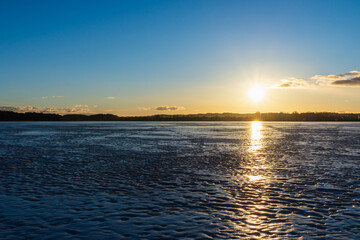Winter landscape with frozen lake at sunrise or sunset. Lake glistening ice reflect a sun.Forest in the background.