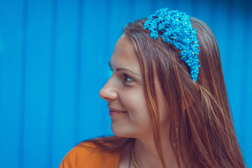 Smiling young woman wearing a decorative floral headband is looking aside. Close-up portrait of a natural girl without makeup over a blue background.