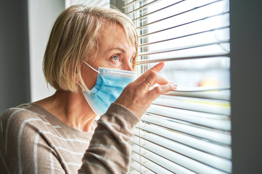 Quarantined senior woman wearing mask looks through blinds