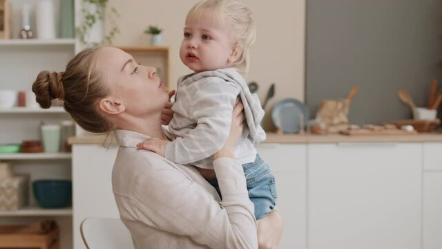 Slow motion of young tired Caucasian female parent sitting at home, picking up upset tear-stained two-year-old son, calming him down