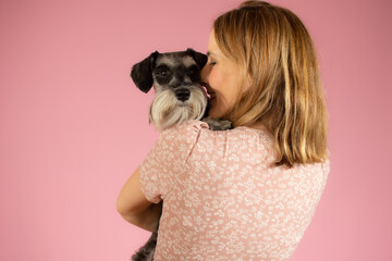 Cute young woman hugs her puppy schnauzer dog. Love between owner and dog isolated over pink background. Studio portrait.
