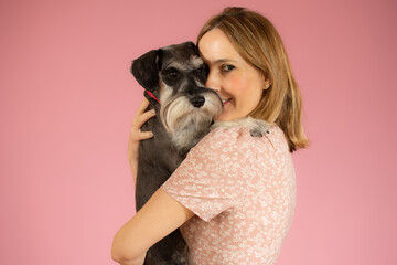 Cute young woman hugs her puppy schnauzer dog. Love between owner and dog isolated over pink background. Studio portrait.