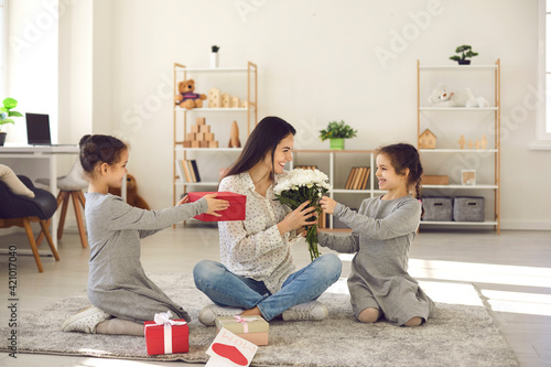 Love and pleasant surprises from kids: Cute little daughters giving their mom presents and flowers on her birthday or Mother's Day. Happy young woman thanking children for wonderful gifts and bouquet
