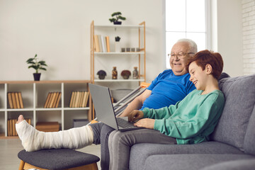 Senior man with injury spends recovery period at home with family. Happy grandson and his grandfather with broken leg in plaster cast sitting on sofa and watching funny movie on laptop computer