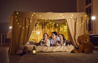 Girls having fun together with mommy. Happy mom and kids enjoying family evening at home. Mother and children lying in cosy tent decorated with fairy lights in dark nursery room with toys and lantern