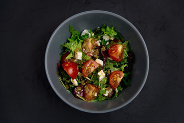 Fresh salad with tomatoes, arugula and cheese cubes on black plate on Dark grey black slate background