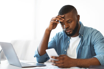 Concerned black freelancer guy looking at smartphone while working at home office