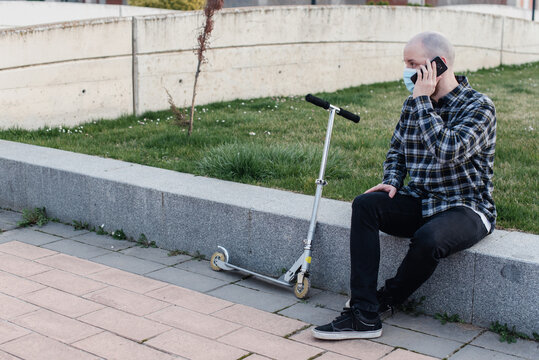 Hombre joven sentado hablando por teléfono mientras lleva una mascarilla desechable quirúrgica.  Hombre con camisa de cuadros con un teléfono móvil al lado de un patinete no eléctrico de metal.