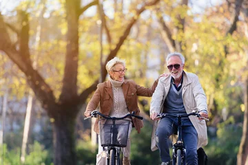Plexiglas foto achterwand Cheerful active senior couple with bicycle in public park together having fun. Perfect activities for elderly people. Happy mature couple riding bicycles in park © lordn