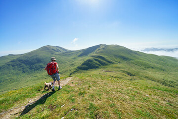 Fototapeta premium A female hiker and their dog walking down from the mountain summit of Meall Corranaich with Ben Lawers and Beinn Ghlas in the distance in the Scottish Highlands, UK landscape.