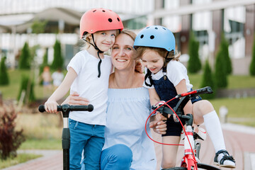 Naklejka premium Mother with two chherful daughters having fun. Woman and girls in helmets riding on bicycle and scooter