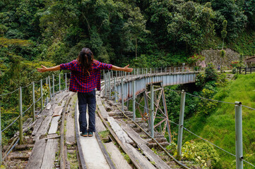 Man from behind with dreadlocks and square shirt standing on the train tracks on a bridge with open...