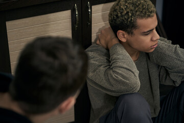 Staged photo illustrates problems and conflicts in gay couple relationships. Wistful and upset swarthy man in gray cardigan is sitting near kitchen cabinet. Other person's head is in the frame.