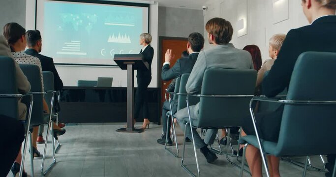 Young beautiful business woman in stylish suit with heels demonstrates her project presentation answering questions from hall.