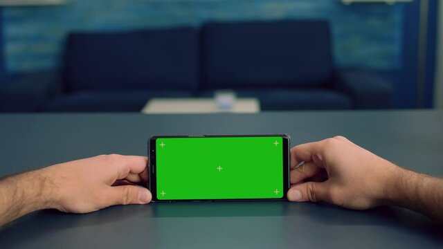 Pov Of Man Hands Holding In Horizontal Mode Smartphone With Mock Up Green Screen Chroma Key Display Standing On Desk In Office Studio. Freelancer Using Isolated Phone For Browsing Social Networks