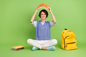 Portrait of attractive funny cheerful girl sitting lotus position holding book over head like roof...