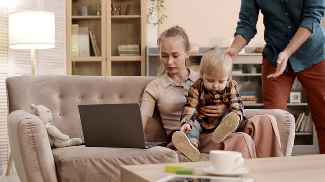 Medium long slow motion of young Caucasian woman sitting on couch with son on her lap, working on laptop computer, father approaching them, taking toddler away