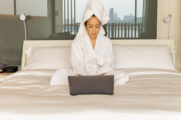 Woman in dressing gown and towel on her head sits in bedroom on bed working on laptop.