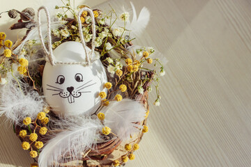 Cute handmade egg in a wreath with bunny ears lying in the nest made of natural materials (wood, branches, grass, flowers, feather) with light wooden background. Easter holiday concept. Top view, copy