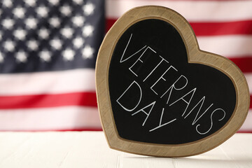 Heart shaped blackboard with phrase Veterans Day on white wooden table against American flag, space...