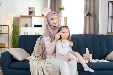 Young Arabian mom in headscarf holds her little pretty girl child on her knees, sitting on the sofa...