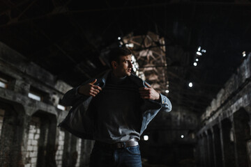 Fototapeta na wymiar Stylish brutal man in jeans jacket standing in the dark old building and looking to the side