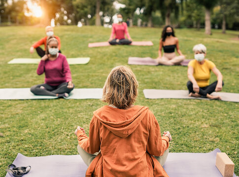 Yoga teacher doing exercise at nature park with multiracial women while wearing safety face mask for coronavirus outbreak - Multi generation people sitting with social distance