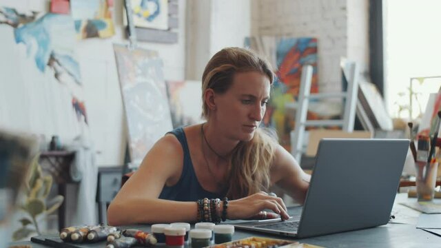 Young Caucasian woman using laptop at desk while working in art studio with paintings in background