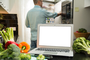 Man using laptop and cooking in kitchen at home. Distant work. Fresh vegetables on kitchen counter. Healthy lifestyle and social media concept. White empty screen