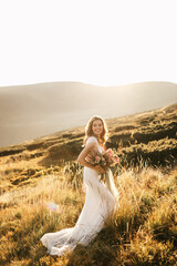 Fototapeta na wymiar Beautiful bride in a white wedding dress stands posing and smiles in the mountains with a bouquet