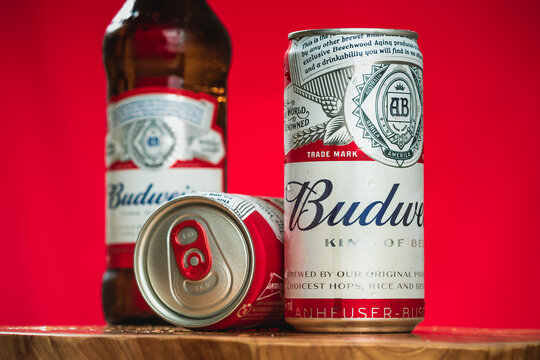 Cerveja Budweiser. Latas e uma garrafa de cerveja da marca Budweiser sobre uma tábua de madeira com plano de fundo em cor vermelha. Cervejaria, Cerveja, Bud, Ambev, Inbev.
