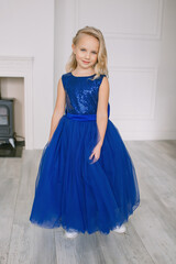 Little beautiful girl in a blue dress stands in a room near the fireplace light interior