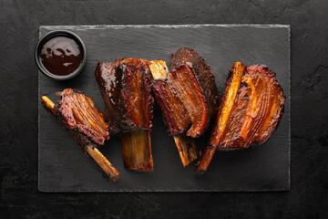 Smoked beef ribs with bone and sauce in a bowl on the side on a black slate board, top view.