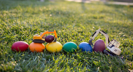 2 metal toy excavator models stand on rainbow-colored eggs on the grass. Business concept of Easter...