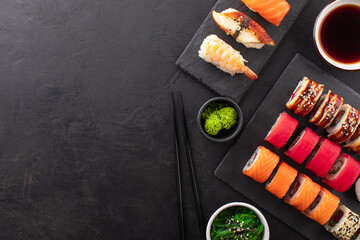 Assorted sushi roll set served on a black slate board, seaweed salad, soy sauce in a bowl and wasabi on the side. Asian food composition on black background, top view, copy space