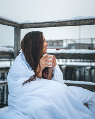 A beautiful woman covered in a blanket sitting on the winter terrace and drinking coffee.