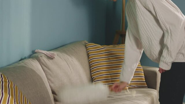 Pretty girl in white sweater in spacious bright room color of sea wave, with window, during cleaning finds dirty sock and hides it under pillow. Medium shot.