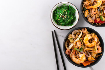 Stir fry noodles with shrimps and mussels in a black bowl and seaweed salad on a white stone background, top view, copy space.
