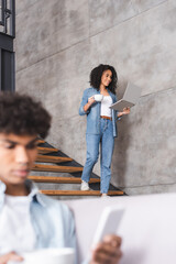 Smiling african american woman with laptop and cup walking on stairs near boyfriend with smartphone