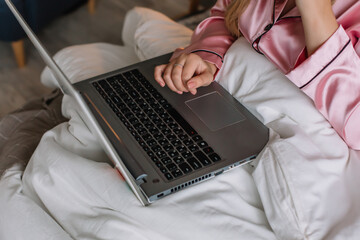 Young woman sits in the bedroom on the bed Dressed pink pajamas on her body Holds a laptop in her hands Female hands on the keyboard