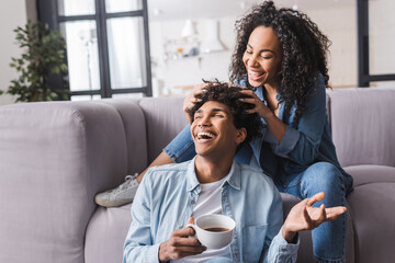 Happy african american woman touching hair of boyfriend with cup at home