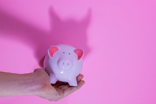 Piggy bank in a hand casting a sinister shadow with devil horns on a pink background, copy space