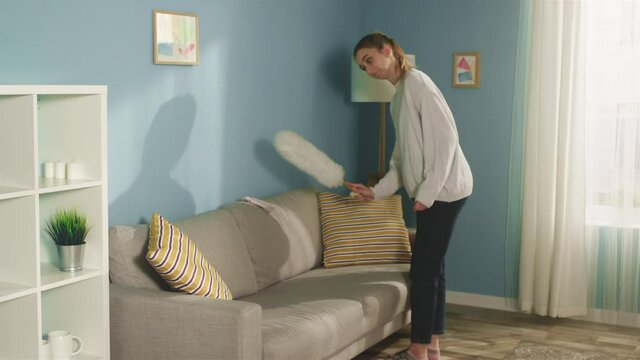 Pretty lady in white sweater in spacious bright room color of sea wave, with window, during cleaning finds dirty sock and hides it under pillow. Long shot.